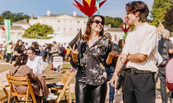Two young adults engage in conversation at an outdoor festival, surrounded by colorful decorations and attendees enjoying the event.